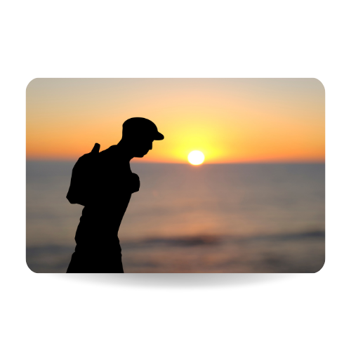 Closeup of a paper cutout in the shape of a young man, wearing a flat cap and carrying a backpack, in front of the ocean at d